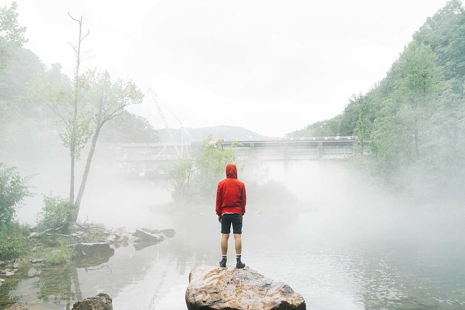 man standing on rock facing foggy creek, person in red hoodie standing on gray rock surround by body of water