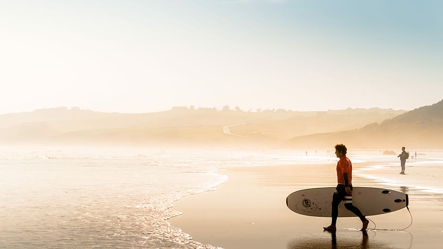 man holding white surfboard walk through ocean under blue sky HD wallpaper