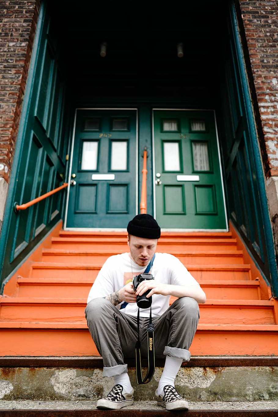 man seating in front of stair while holding DSLR camera, man sitting on stairs in front of green doors during daytime