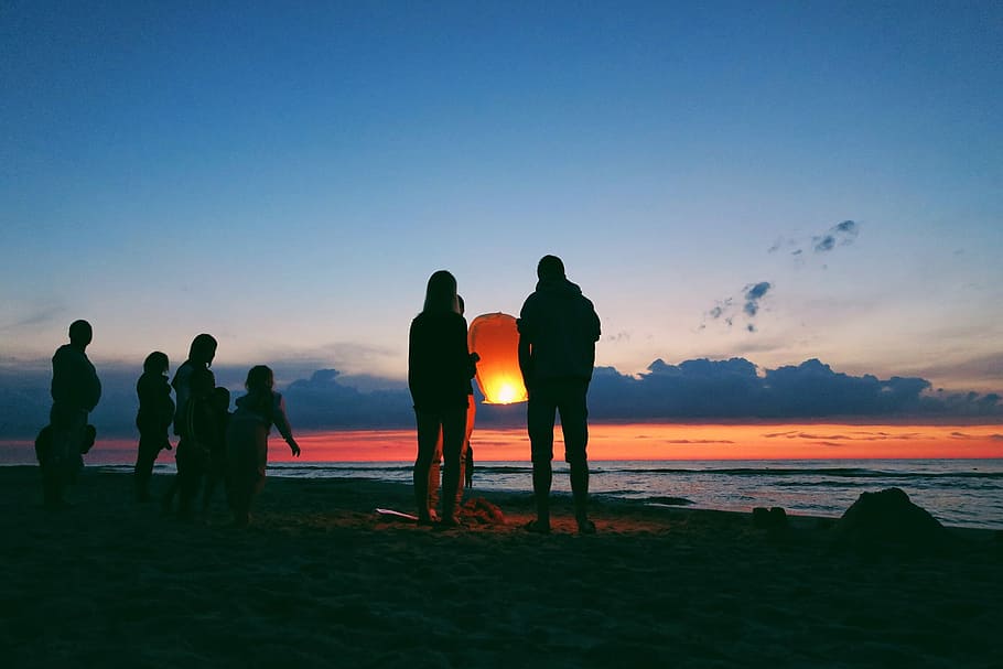 silhouette of person holding sky lantern, silhouette of people on seashore at golden hour HD wallpaper