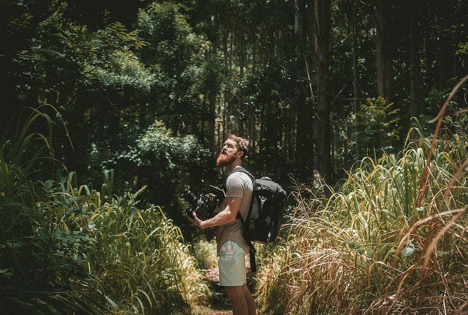man holding camera in the forest, man wearing brown shirt with black backpack
