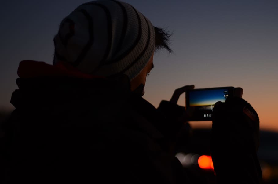 man holding phone to take a picture, person taking photo wearing hat during nighttime HD wallpaper