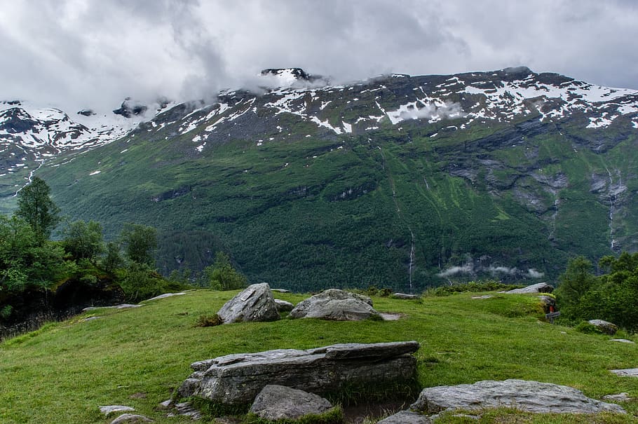 gray and black rocks surrounded by green grass near mountain under white sky at daytime, green mountain covered with snow during daytime HD wallpaper