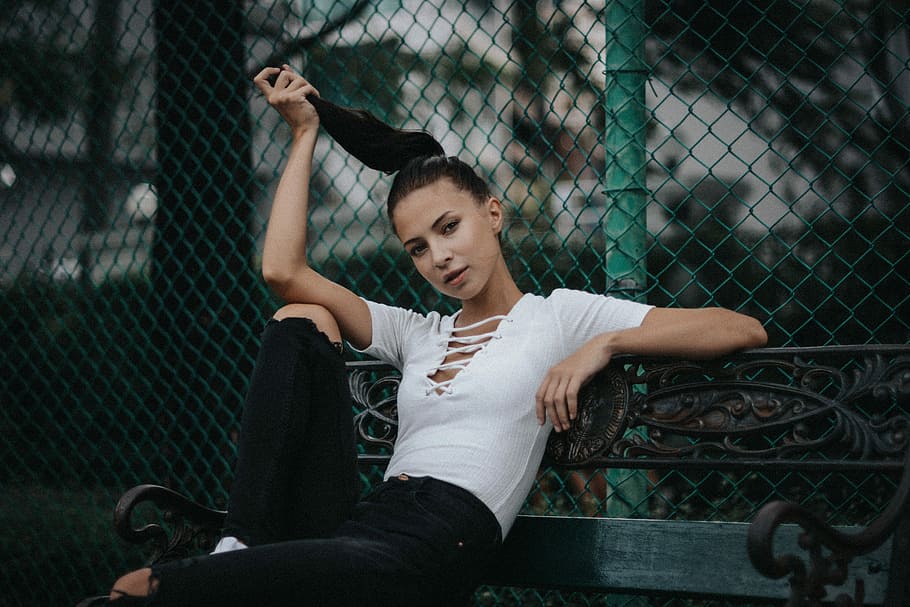 woman wearing white cap-sleeved crop top and distressed black denim jeans sitting on black metal bench beside chain-link fence during daytime, woman in black jeans sitting in bench, HD wallpaper