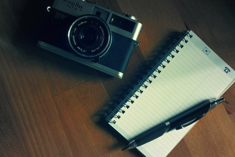 flat lay photography of grey and black Minolta MILC camera beside black lined notebook and retractable pen on wooden table
