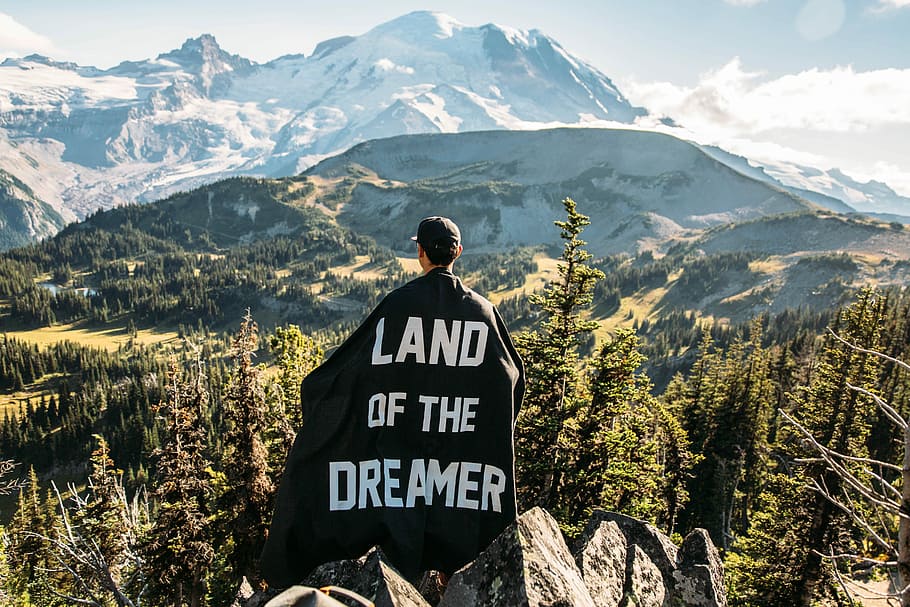person standing on mountain peak with black land of the dreamer-printed cape on his back, man wearing black and of the dreamer printed cape while standing on gray mountain overlooking forest with green trees at daytime