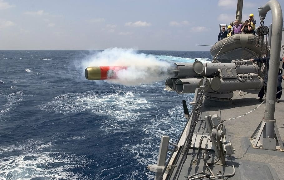 gray torpedo gun launches torpedo towards sea with watching men at distance at day