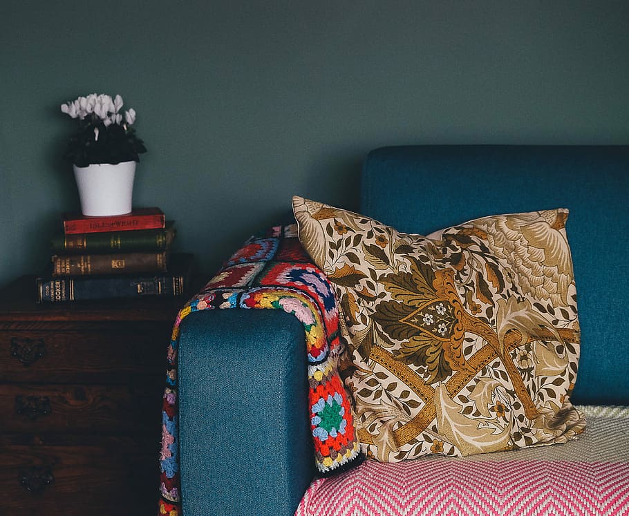 yellow throw pillow on blue couch beside on pile of books on top of drawer, brown and orange throw pillow on top of teal velvet sofa