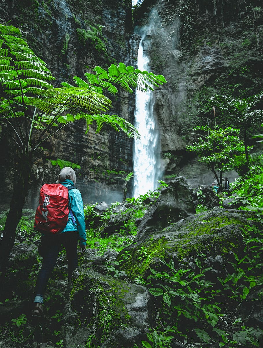 person walking towards waterfall at daytime, person wearing blue windbreaker jacket and black pants carrying red backpack near waterfalls