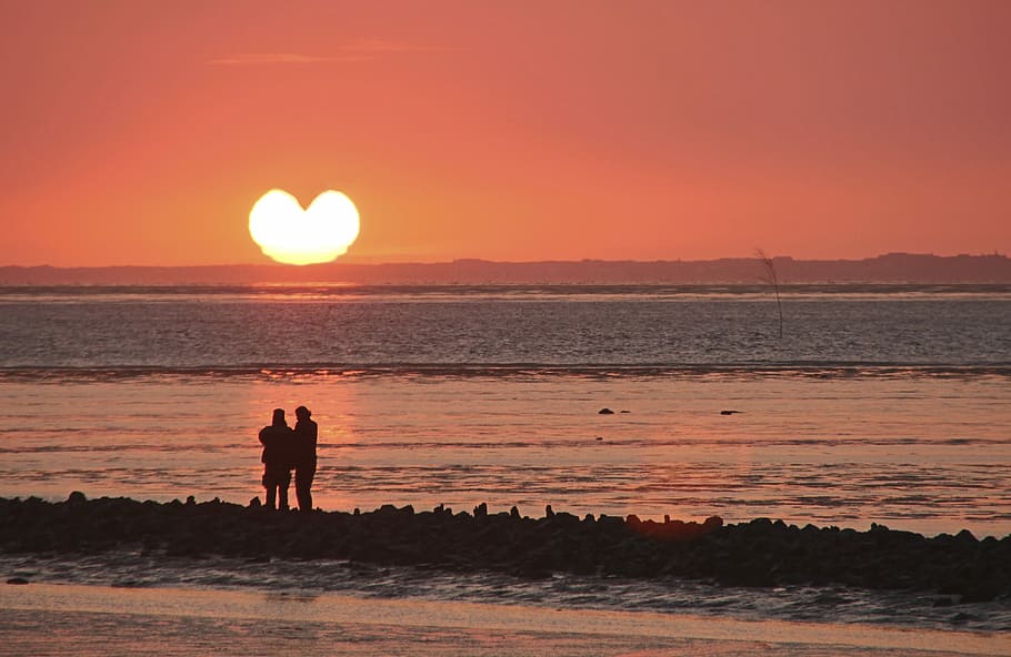 silhouette photo of couple standing near shore facing heart sun