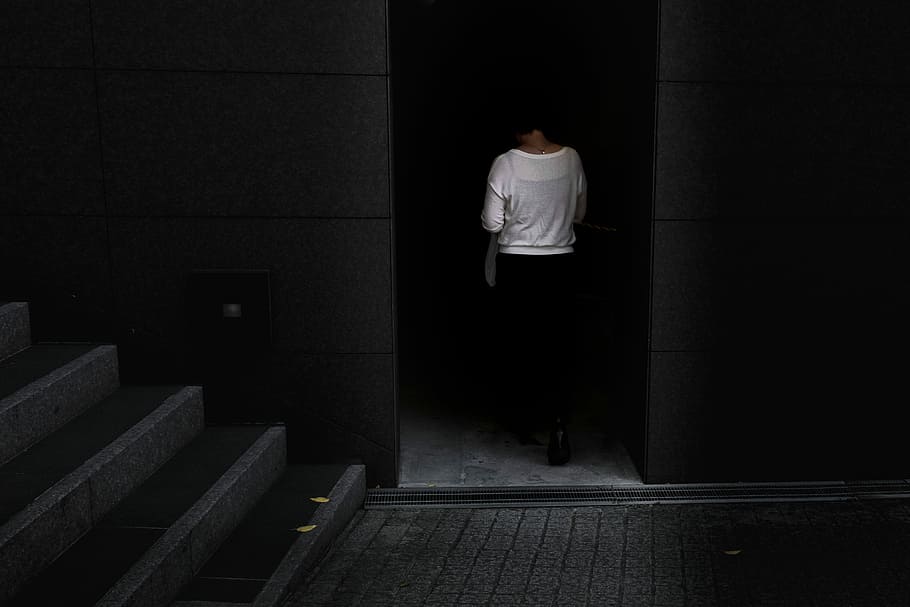woman walking near gray stair inside building, woman in white top walking through the dark
