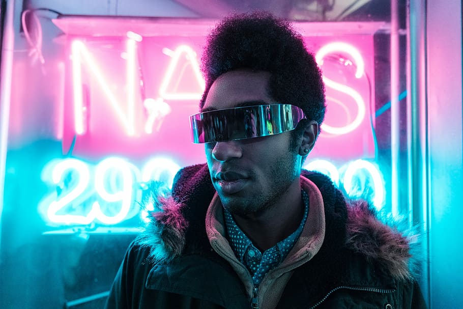man wearing black parka jacket and black sunglasses in front of neon signage, person wearing sunglasses standing near neon sign