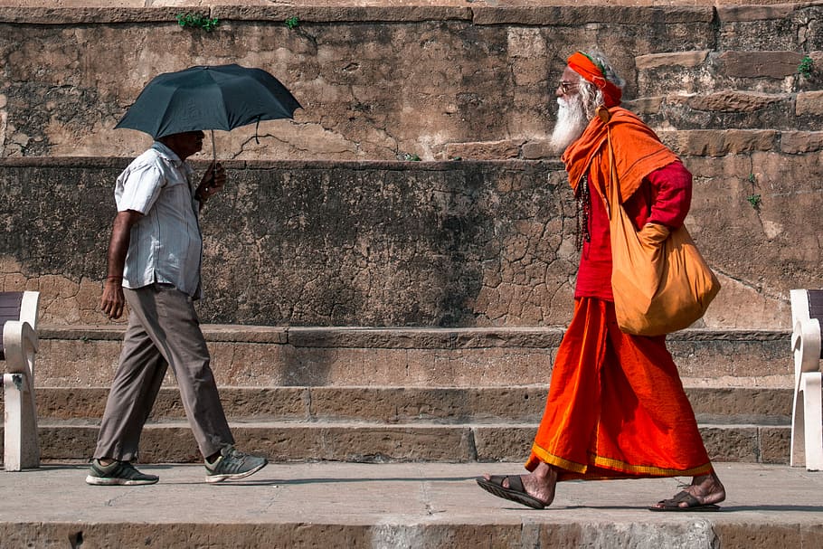 man walking beside man holding umbrella, man in orange and red dress walking towards man holding umbrella during daytime HD wallpaper