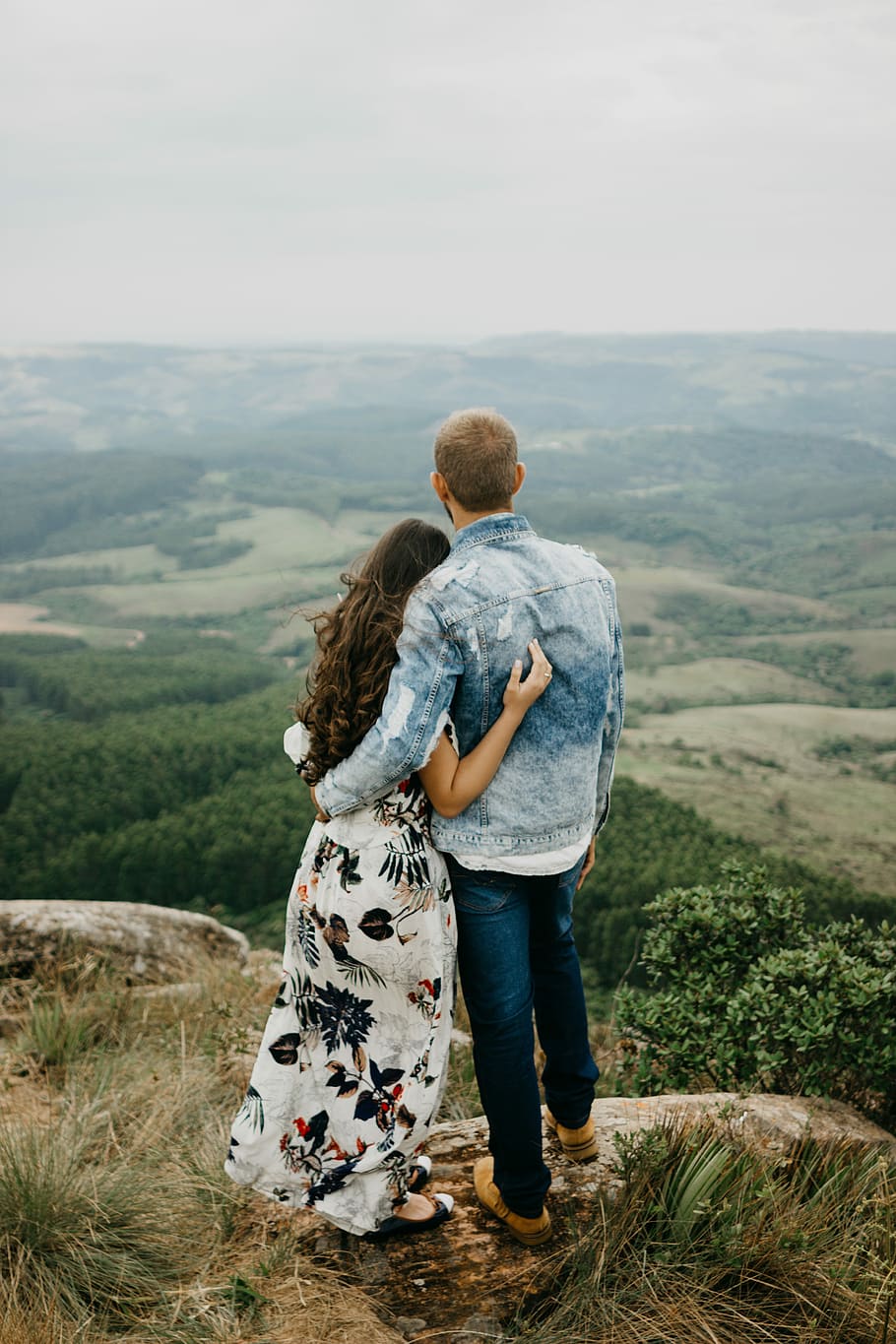 man and woman standing on cliff during daytime, man in blue denim jacket beside woman in black and white floral dress standing in front of mountains during daytime