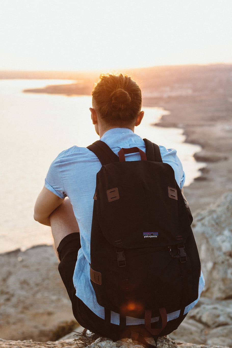 person in black knapsack sitting on cliff, tilt-shift lens photography of person sitting on rock surface facing body of water during daytime HD wallpaper