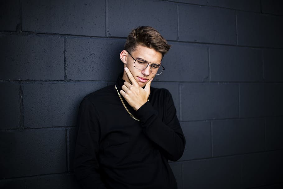 man leaning on wall while looking down wearing eyeglasses and necklace with right hand on chin, man wearing black long-sleeved shirt holding his chin