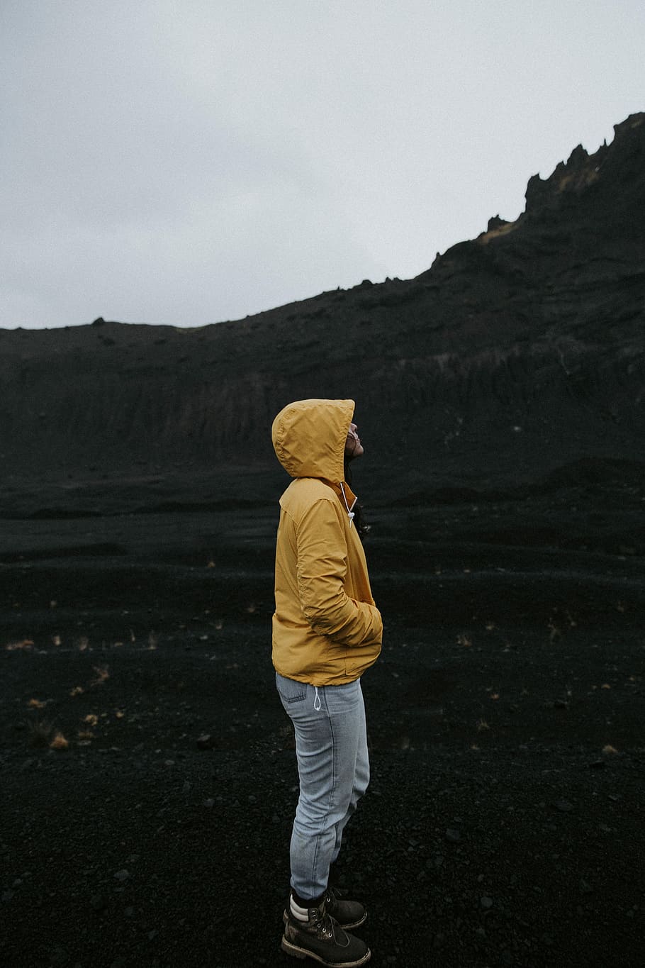 person standing beside mountain, person in yellow hoodie standing on black soil; HD wallpaper