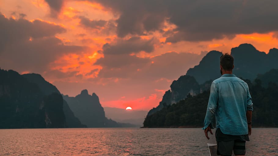 man standing near edge facing body of water during sunset, man wearing blue chambray shirt facing body of water with view of mountains during sunset photo, HD wallpaper