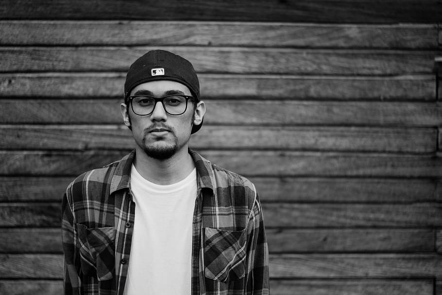 gray scale photo of man wearing glasses, cap, flannel and white shirt, grayscale photo of man behind wall