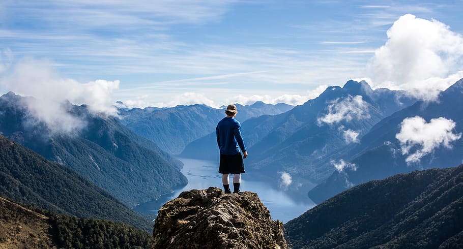 person standing on the top of high rise stone, man standing on rock formation in front of lake surrounded by mountains during daytime