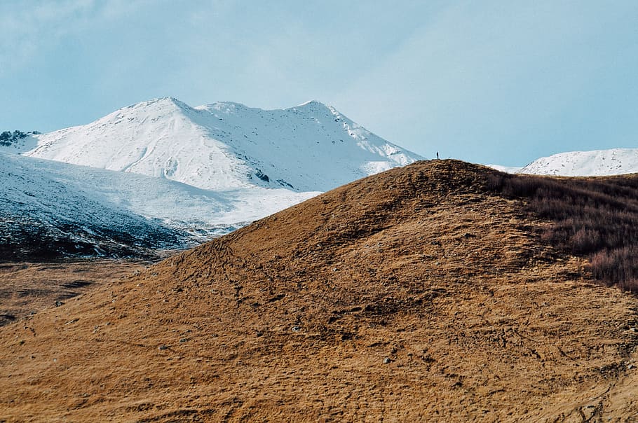 brown mountain near mountain covered with snow, person standing on hill looking to snow covered mountain HD wallpaper