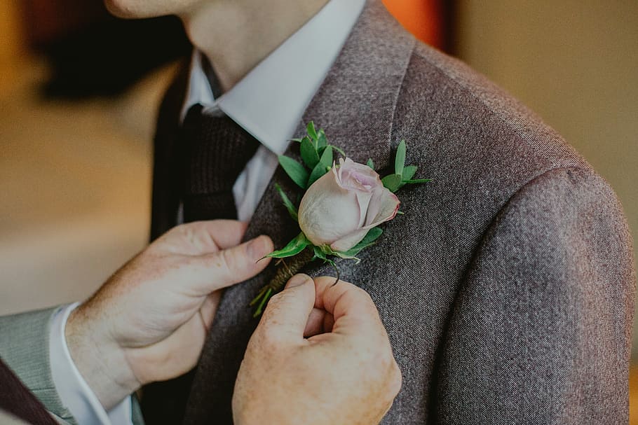 person putting pink rose pin on person's notched lapel suit jacket, man holding brown blazer, HD wallpaper