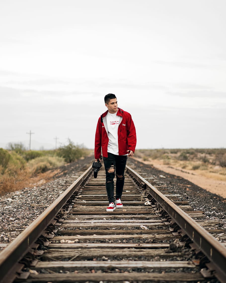 man wearing jacket waking on train rail, man walks on brown train rail during daytime HD wallpaper