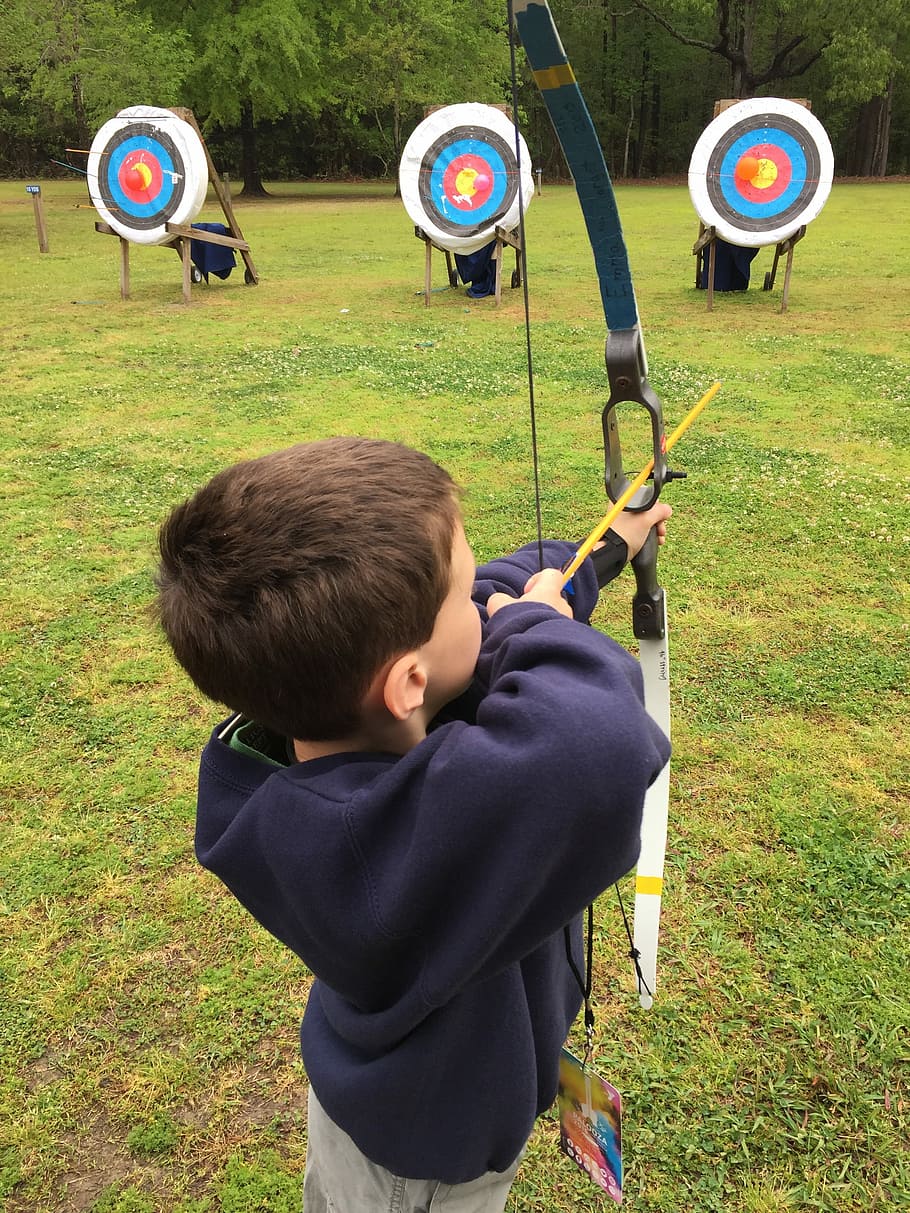 boy using composite bow aiming on target on green grass during daytime