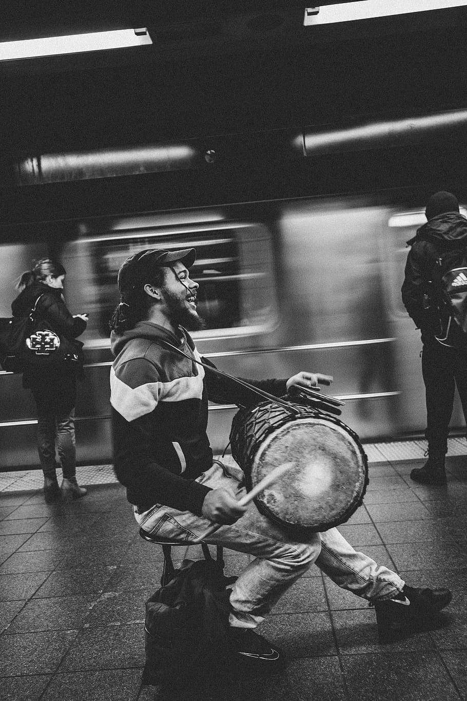 man playing drum in subway, greyscale photography of man playing drum on train station beside two person waiting for train