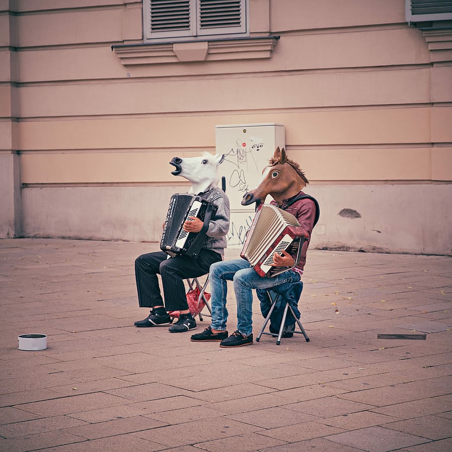 two person wearing horse heads sitting on folding chairs while playing accordions beside brown concrete building, two men wearing horse masks playing accordions while sitting on sidewalk during daytime
