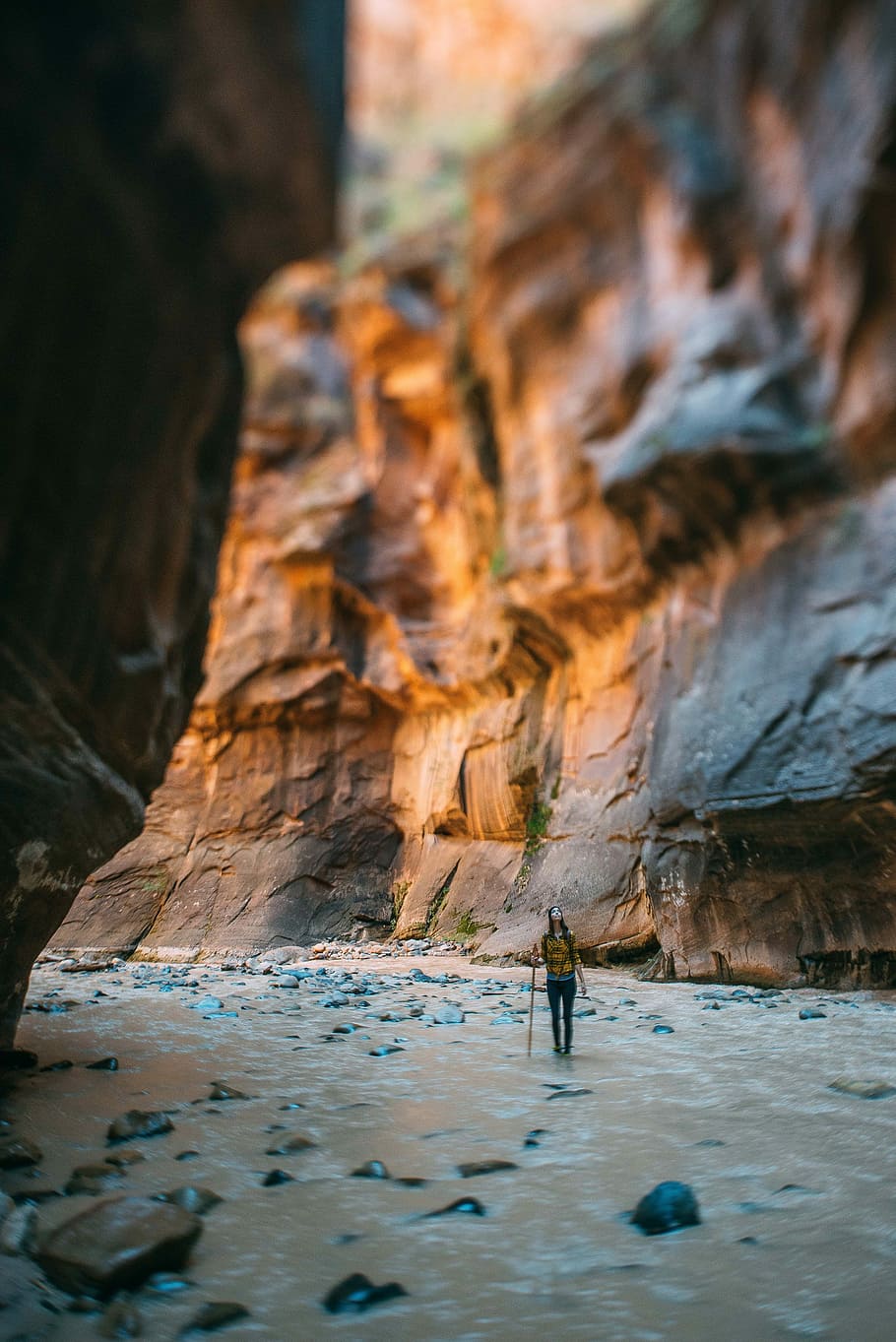 woman holding stick looking upward between cliff during daytime, woman stands on river beside rocks under mountain during daytime HD wallpaper