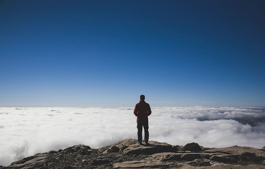 man standing on brown mountains facing sea of clouds, person standing on rock formation near clouds under blue sky during daytime HD wallpaper