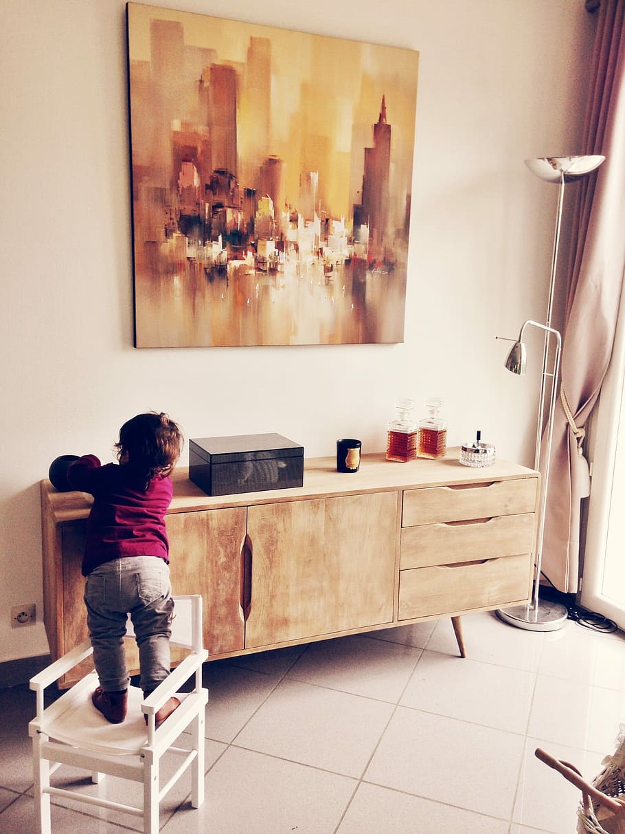 child standing on white wooden armchair in front of wooden sideboard