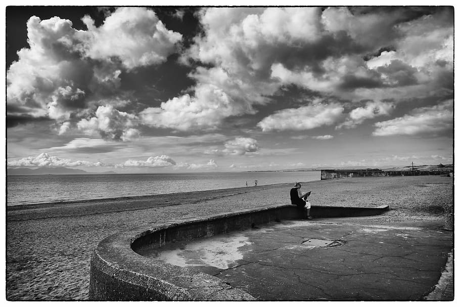grayscale photography of person sitting on concrete barrier near seashore HD wallpaper