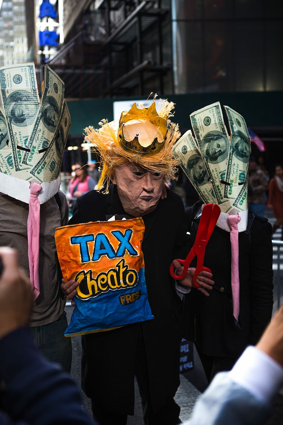 tilt shift lens photo of three men in costumes, person wearing Donald Trump mask with crown holding tax cheato chip bag and red scissors walking on street surround by people HD wallpaper