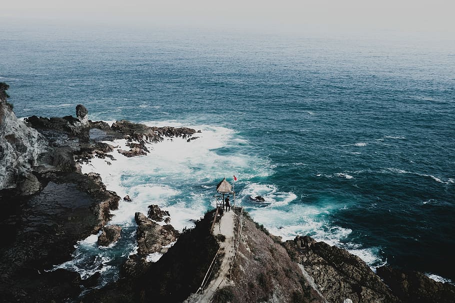 person standing on cliff facing green sea, aerial photography of body of water near island HD wallpaper