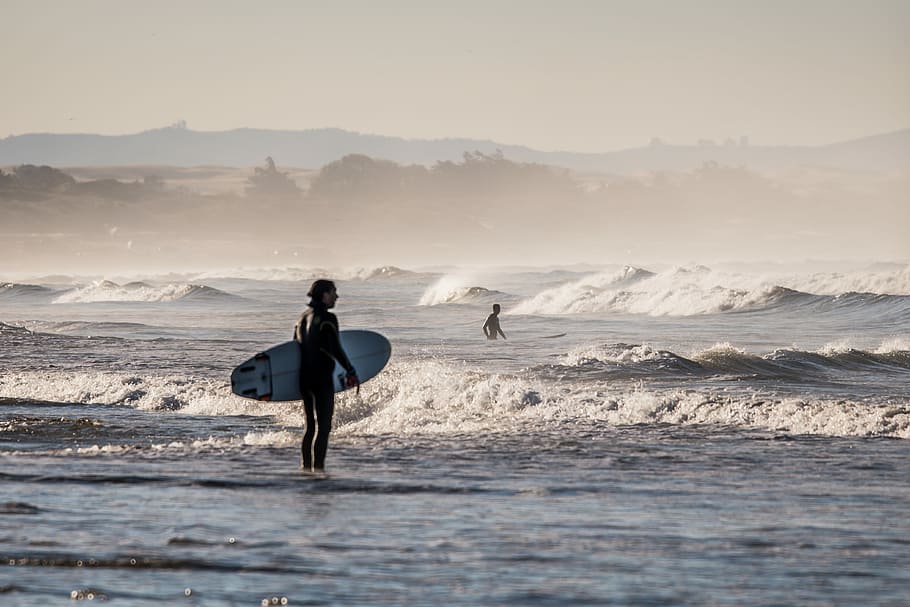 person holding white surfboard standing on body of water under gray sky during daytime, person with surfboard standing on body of water HD wallpaper