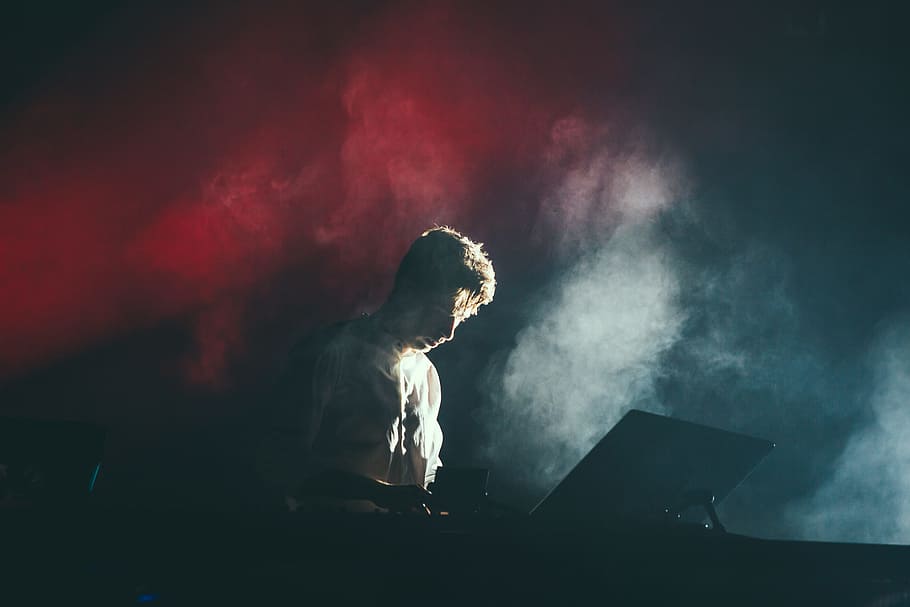 A musician in a white shirt on stage with white and red lights breaking through the smoke behind his back, DJ playing music