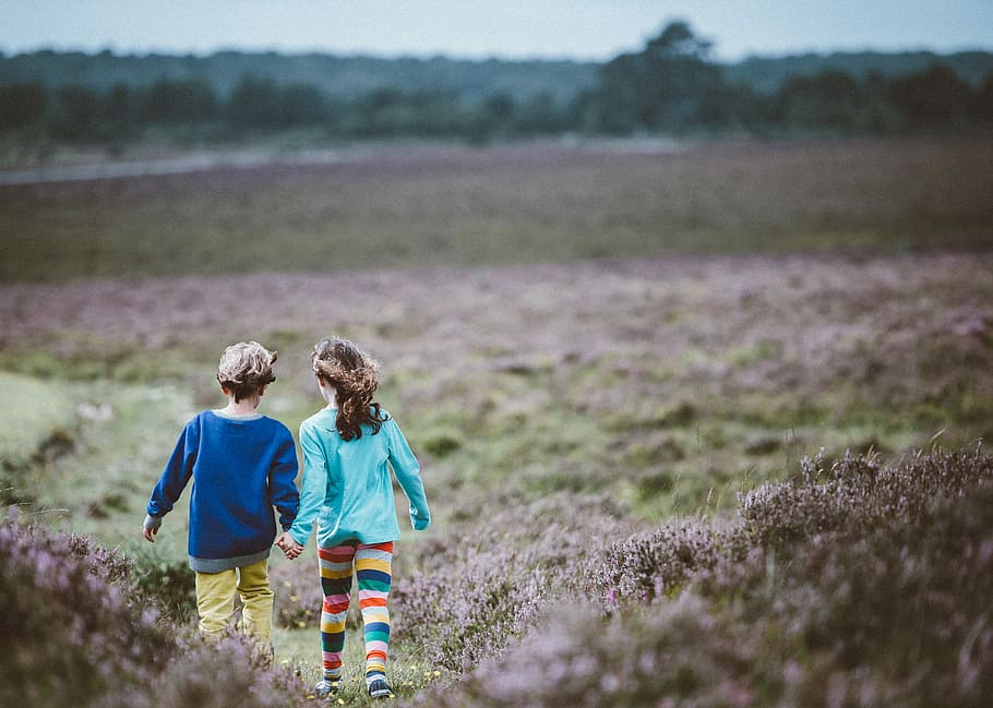 two children holding hands while walking in the middle of the fields, boy and girl holding hands while walking on grass open field during daytime