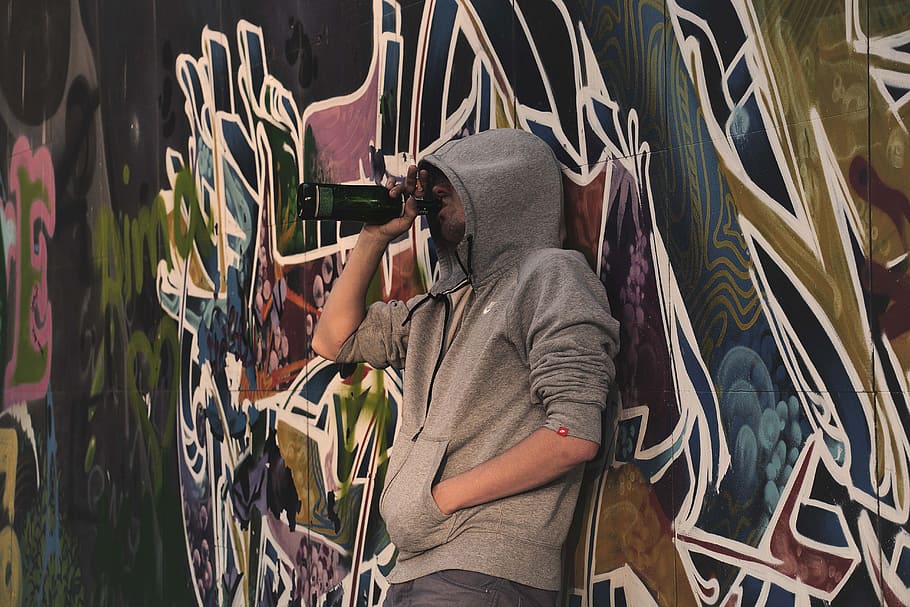 man leaning on graffiti tagged wall while drinking beverage out of bottle