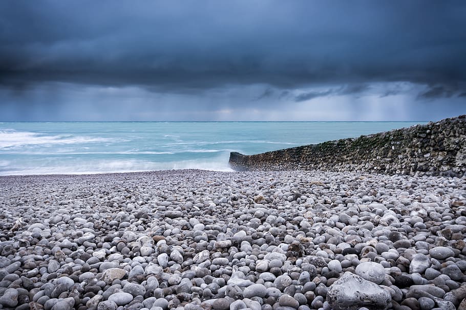 body of water across pebbles under dark sky, body of water near rock wall at daytime HD wallpaper
