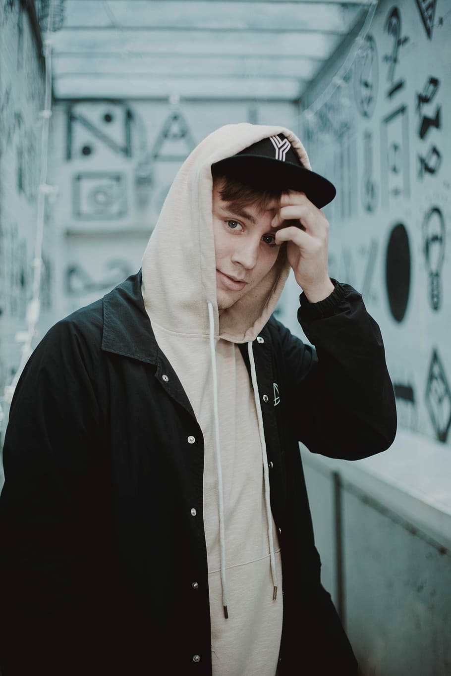man in hoodie standing near white painted wall, man holding his forehead standing near wall HD wallpaper