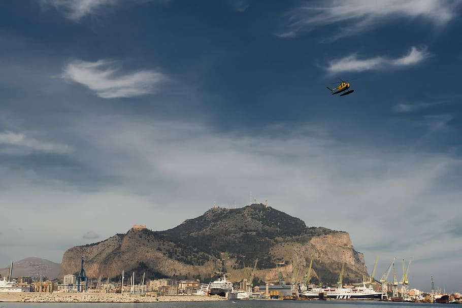 yellow helicopter above assorted-color boats near dock, white boat on dock near mountain during daytime HD wallpaper