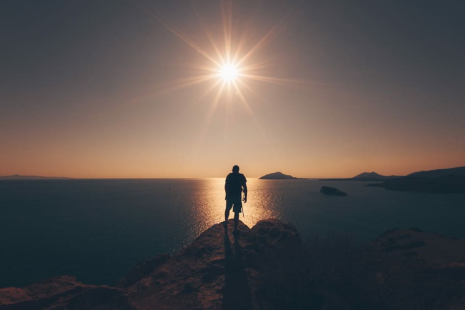 person standing on rock facing sea taken during golden hour, silhouette of man standing on cliff in front of ocean during golden hour HD wallpaper