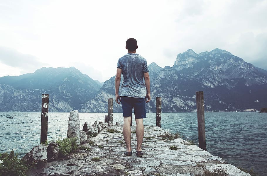 man standing on concrete dock in front of mountain during daytime HD wallpaper