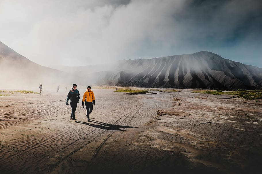 two person standing on brown sands, two man walking near mountain at daytime HD wallpaper
