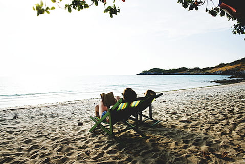 person reading book while lying on lounge chair near ocean during daytime HD wallpaper
