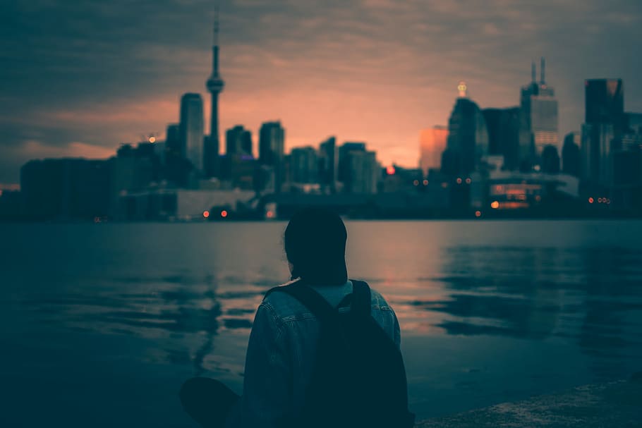 person with backpack sitting near body of water with buildings ahead, man wearing blue denim jacket with black backpack in front blue ocean water HD wallpaper