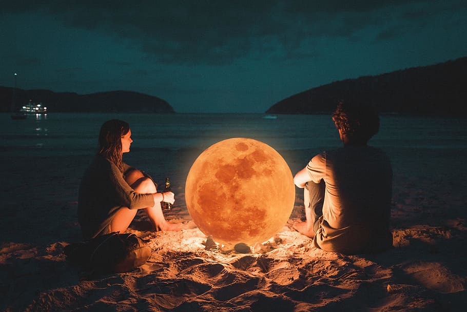 man and woman sitting on sand near sea shore during night time HD wallpaper