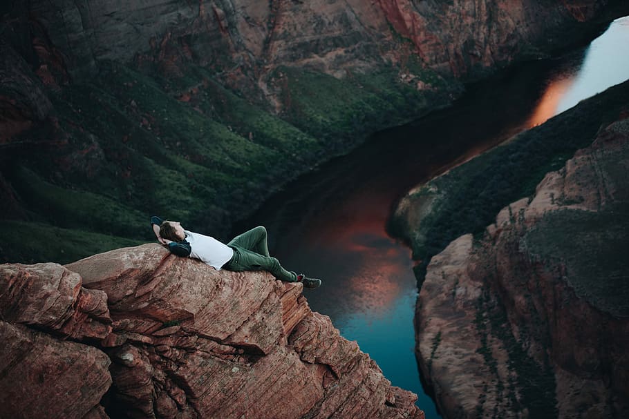 man lying on brown mountain near body of water, person lying on mountain cliff HD wallpaper